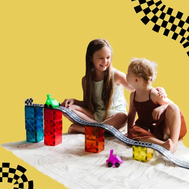 Δύο παιδιά κάθονται στο πάτωμα, χαμογελούν και παίζουν με πολύχρωμα μαγνητικά πλακίδια MAGNA-TILES, αυτοκινητάκια και μια πίστα πάνω σε ένα χαλί με μοτίβα, με φόντο κίτρινο χρώμα και μαύρα καρό σχέδια.