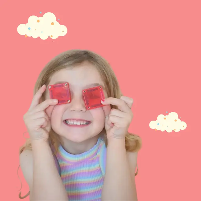 Χαμογελαστό παιδί κρατά δύο κόκκινα ημιδιαφανή τετράγωνα microMAGS πάνω από τα μάτια του, με ροζ φόντο και σύννεφα καρτούν.